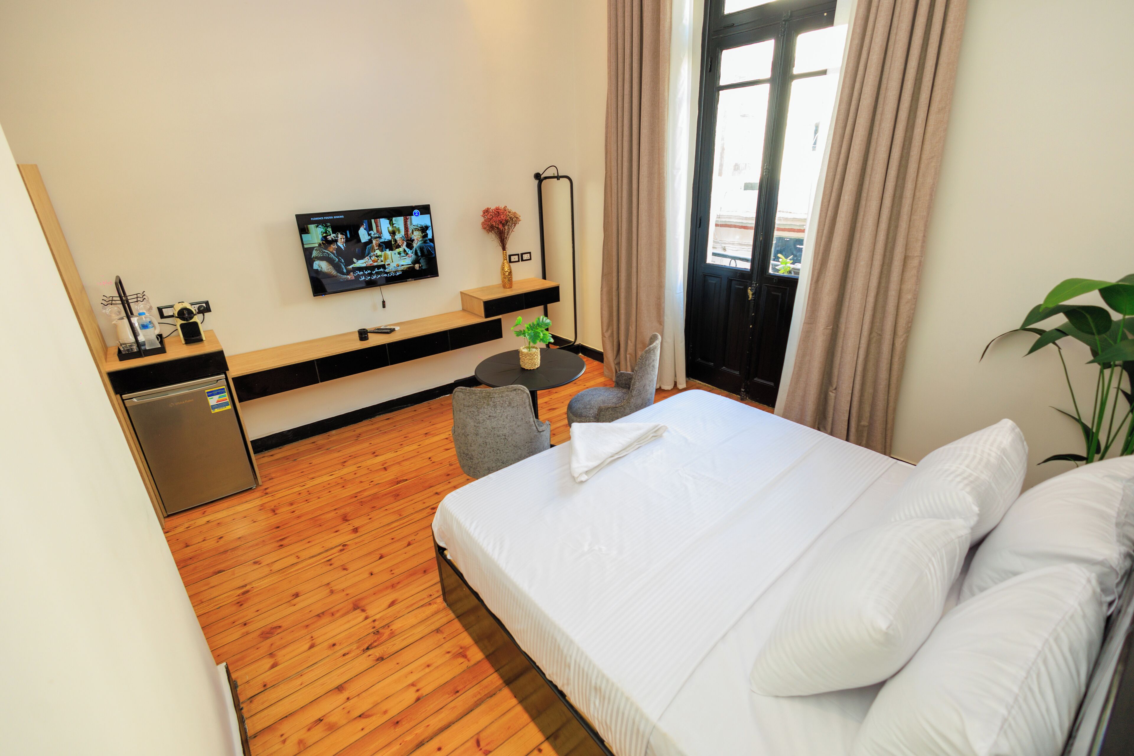 A hotel room with a wooden floor, a bed with white linens, a television on the wall, a small table with a plant, and a door leading to a balcony.