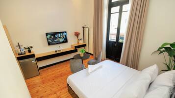 A hotel room with a wooden floor, a bed with white linens, a television on the wall, a small table with a plant, and a door leading to a balcony.