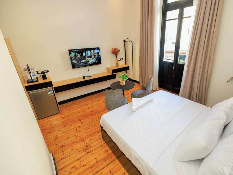 A hotel room with a wooden floor, a bed with white linens, a television on the wall, a small table with a plant, and a door leading to a balcony.