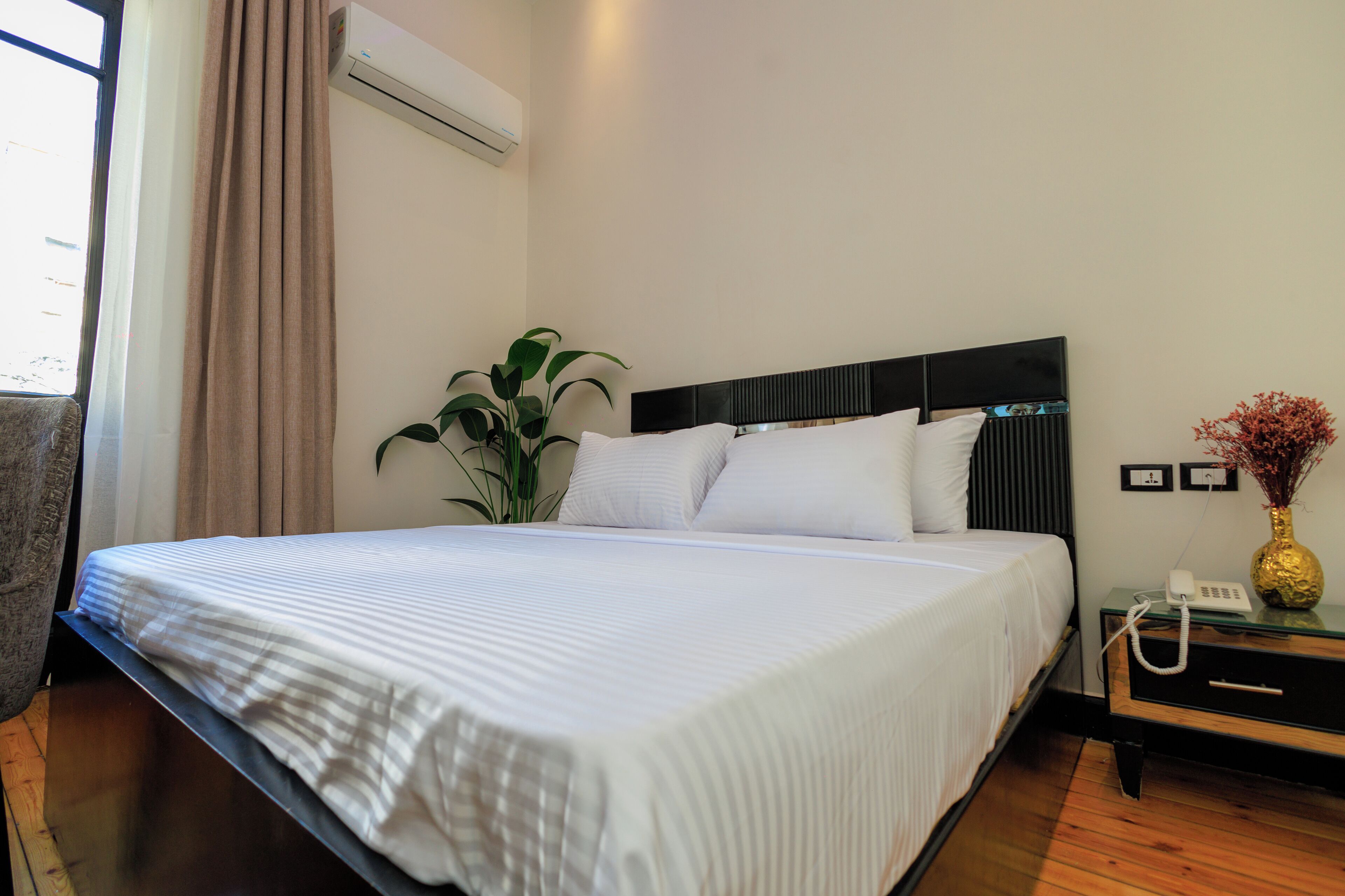 Una cama bien hecha con sábanas blancas, una mesita de noche con un teléfono y un florero, y una planta en maceta al lado de la cama.