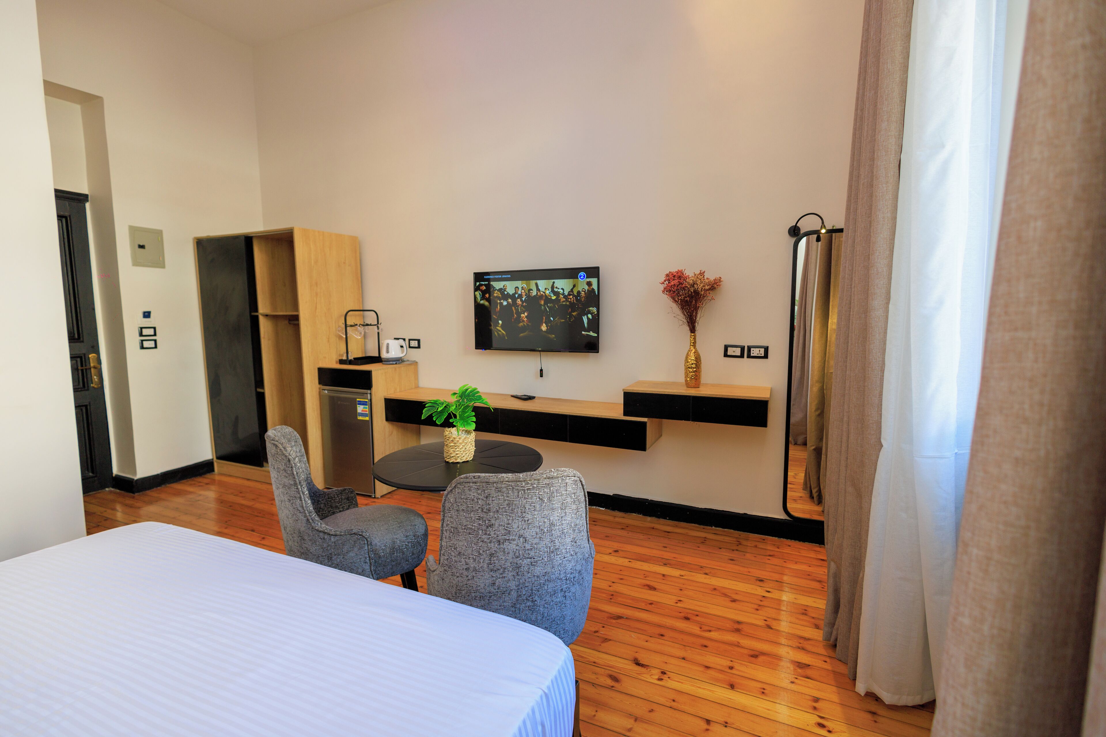 A hotel room with a bed, two armchairs, a small table with a plant, a TV on the wall, and a kitchenette area.