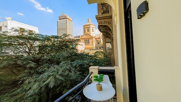 Ein Balkon mit einem Blumentopf, einem kleinen Tisch und Blick auf ein Gebäude mit einer Kuppel und einem Glockenturm.