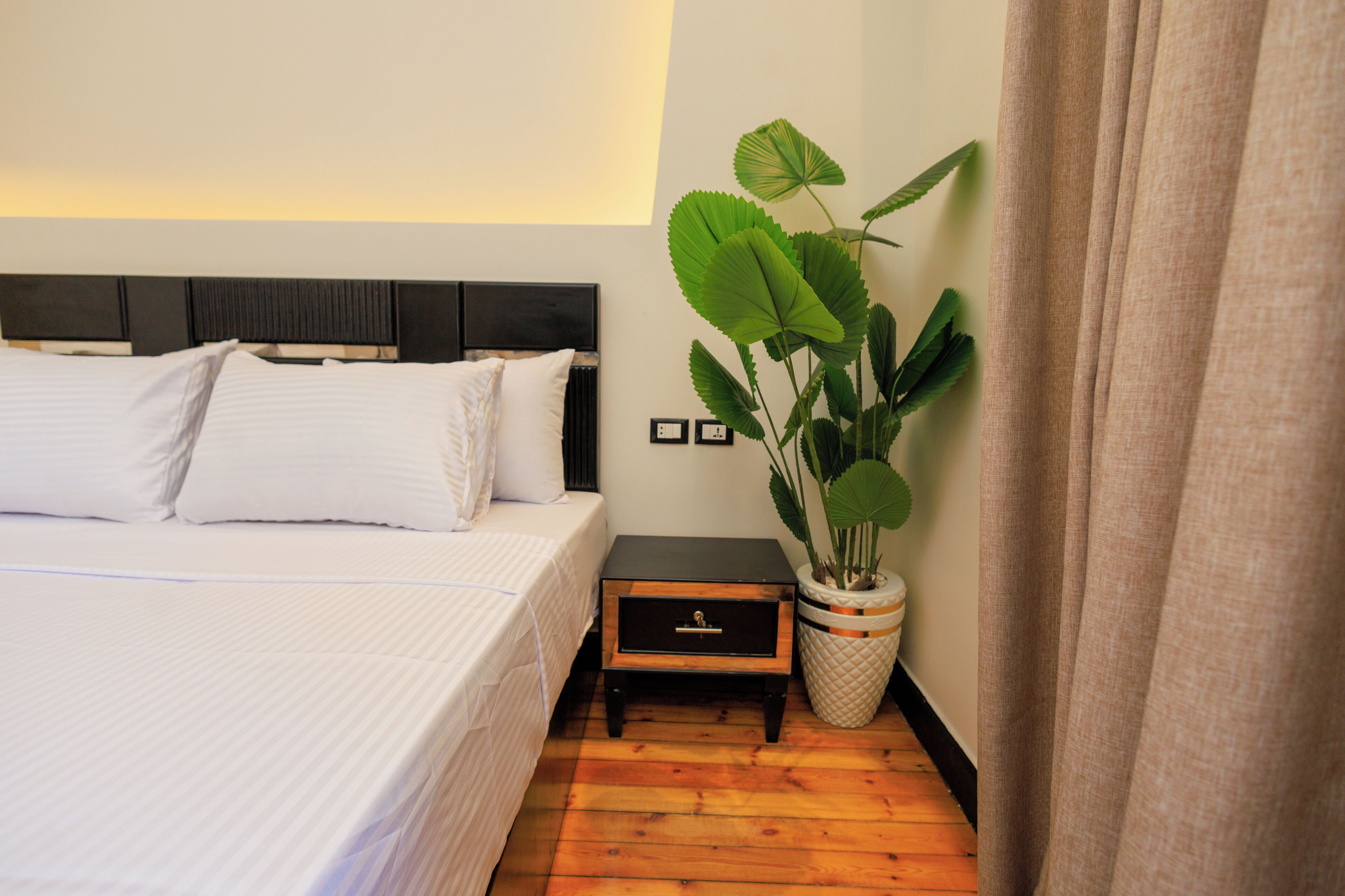 Una cama bien tendida con sábanas blancas, una mesita de noche con una planta y un suelo de madera.