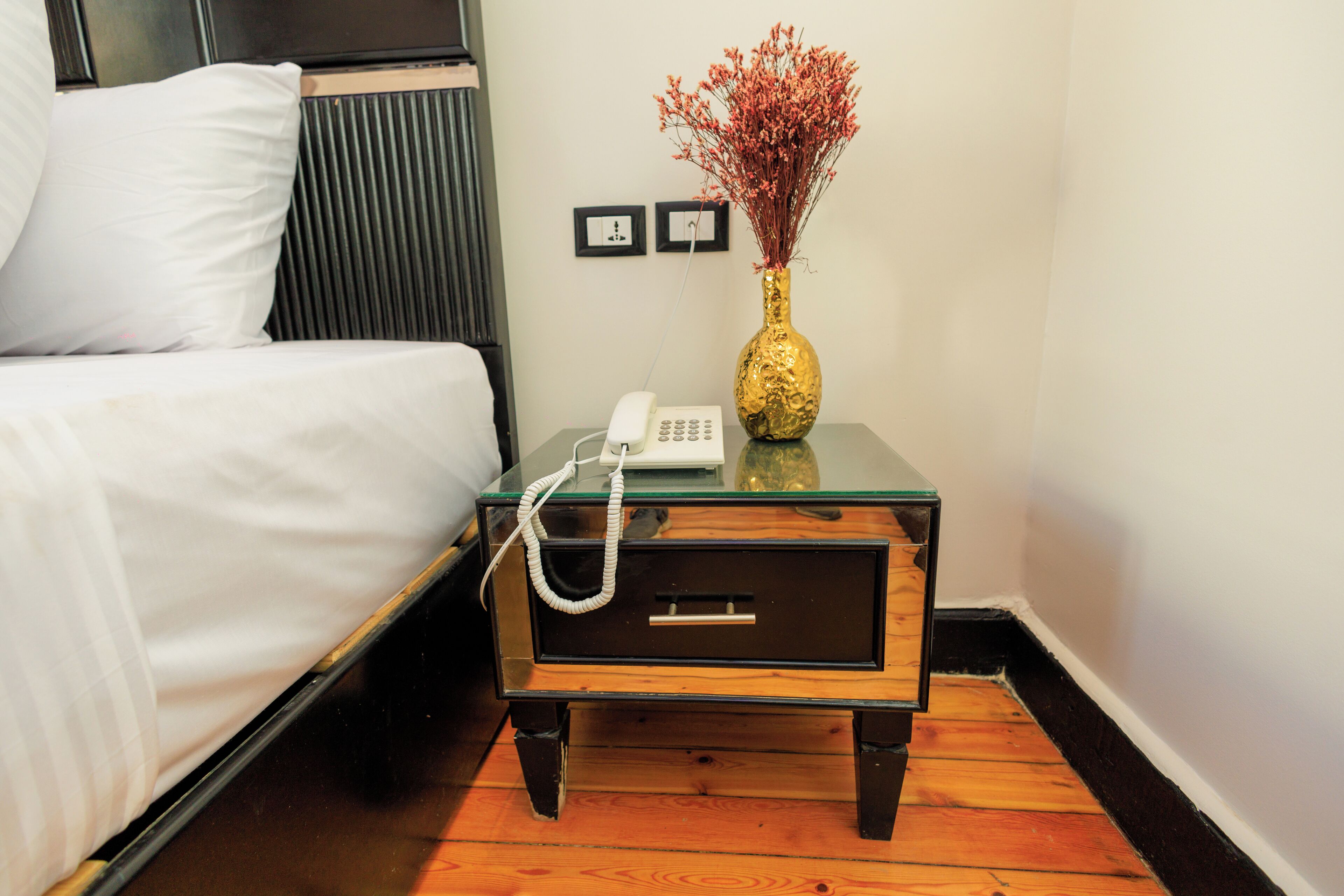 A hotel room with a bed, a bedside table with a telephone, and a vase with dried flowers.