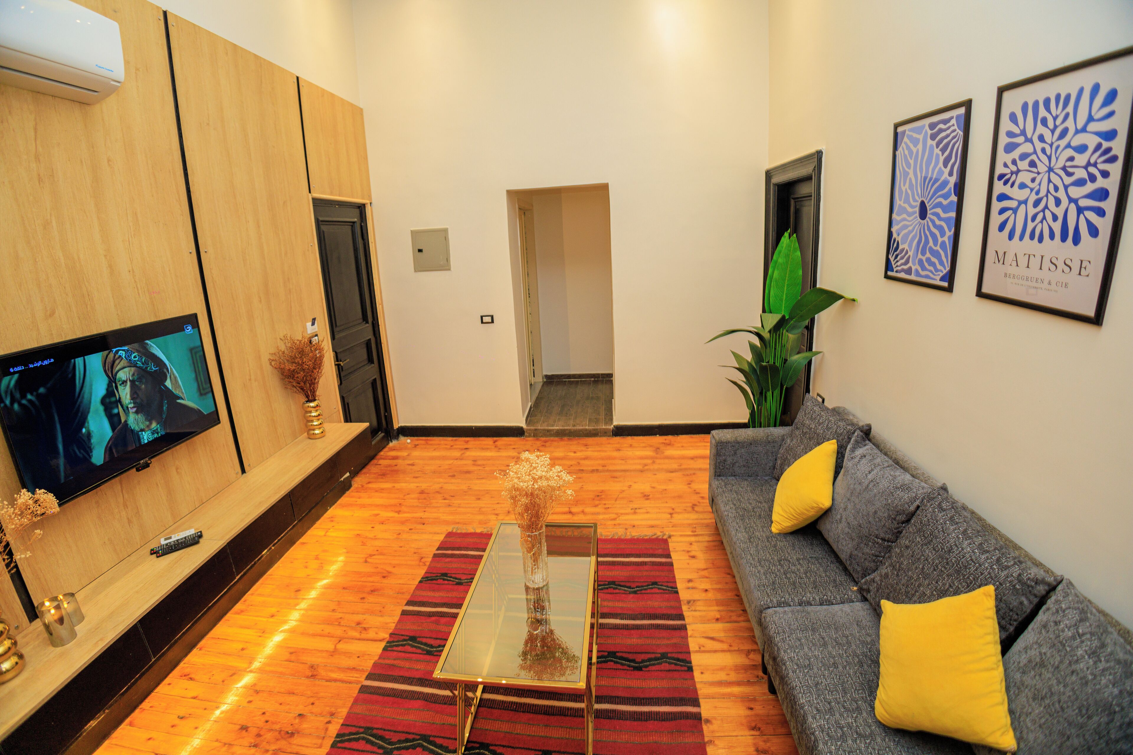 Een moderne woonkamer met een flatscreen televisie, een glazen salontafel, een grijze bank met gele kussens en een rood tapijt met patroon.