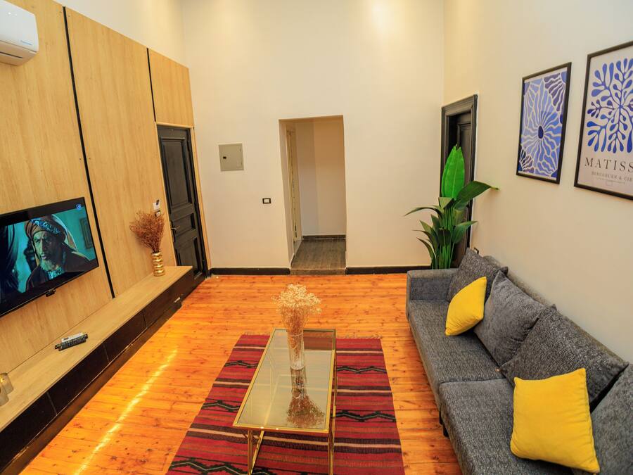 Een moderne woonkamer met een flatscreen televisie, een glazen salontafel, een grijze bank met gele kussens en een rood tapijt met patroon.