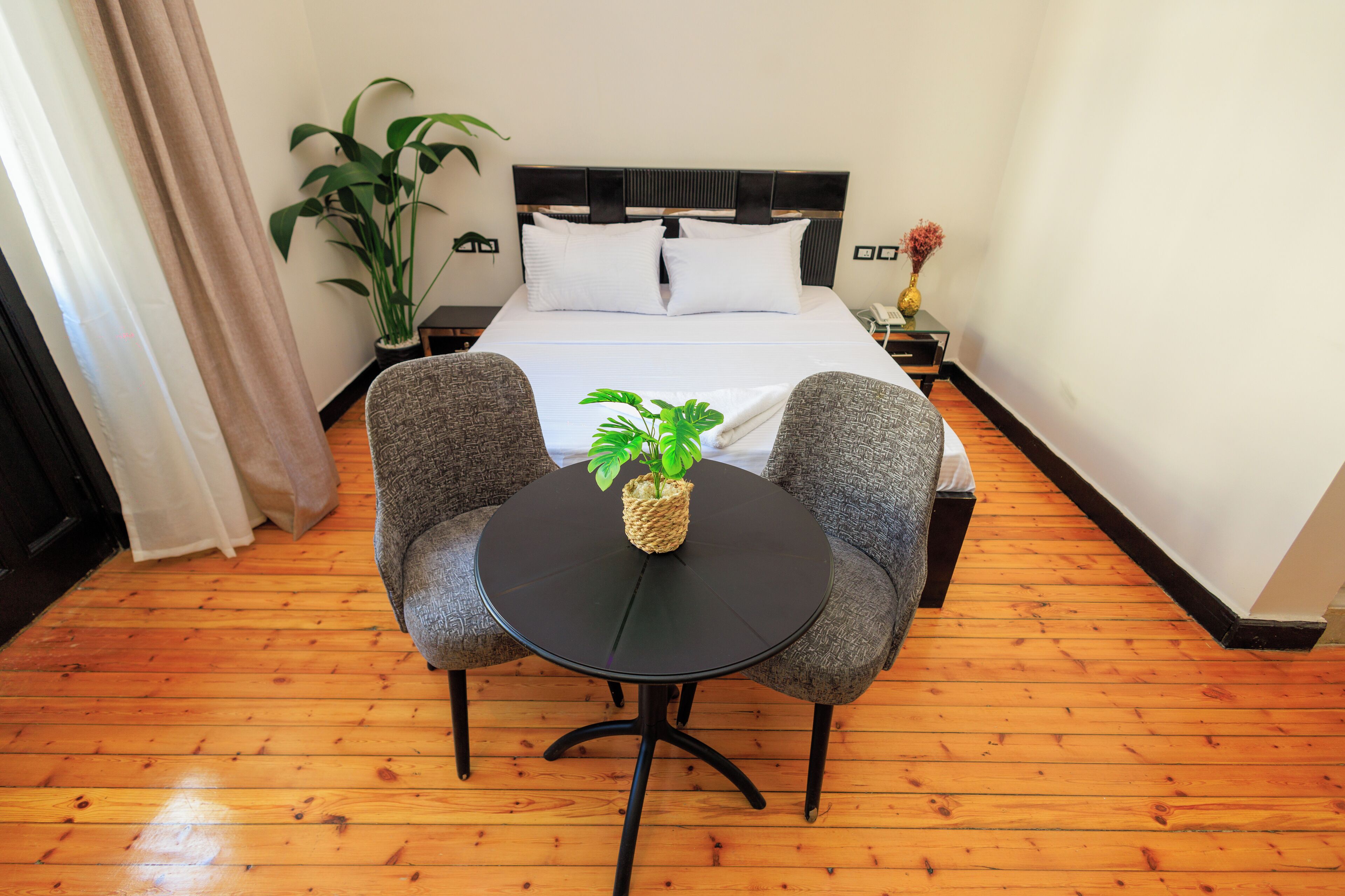 A hotel room with a bed, two armchairs, a small round table with a plant, and a potted plant beside the bed.