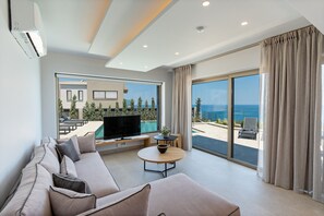 Ein modernes Wohnzimmer mit einer Couch, einem Fernseher, einem runden Tisch und einem großen Fenster mit Blick aufs Meer.