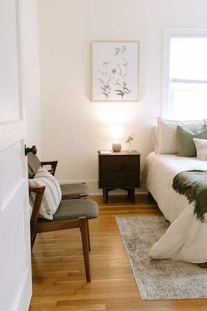 A bedroom with a bed, a nightstand with a lamp, a chair, and a framed botanical print on the wall.