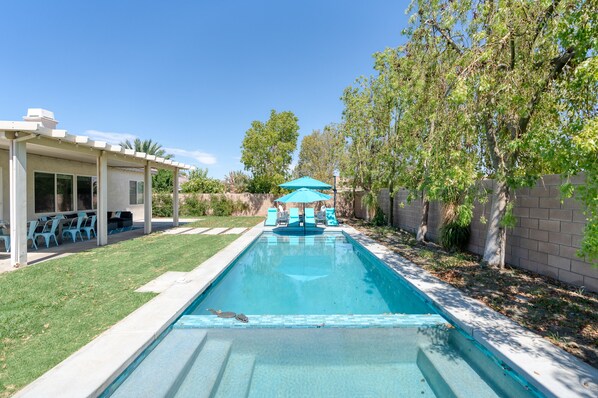 A rectangular swimming pool with a tiled edge, surrounded by a grassy area and a covered patio with blue chairs.
