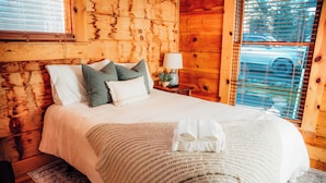 Dormitorio de cabaña de madera con cama, almohadas, lámpara y ventana con persianas.