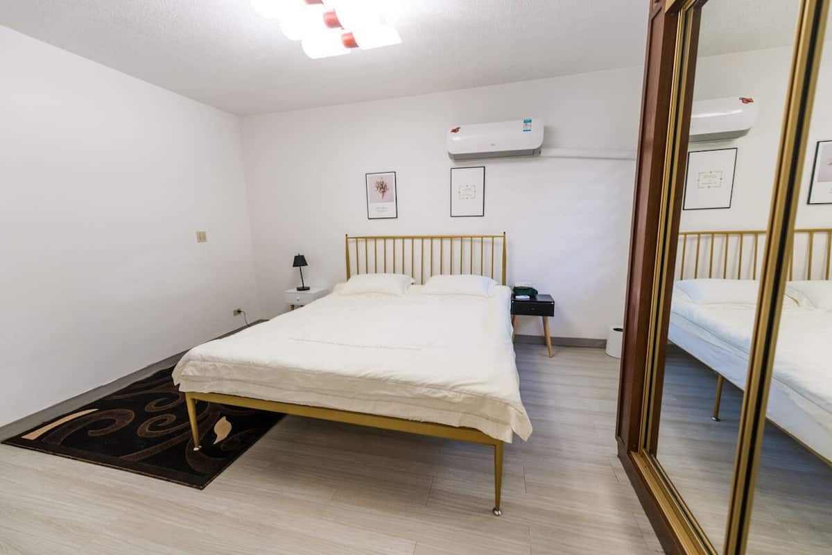A clean, well-lit bedroom with a single bed, a bedside table, a nightstand, an air conditioning unit, and a mirror.