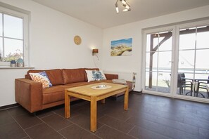 A living room with a brown leather sofa, a wooden table, a wall clock, a painting, and a large sliding glass door leading to an outdoor area. - Chic apartment with terrace, free swimming pool & sauna - Haus De niege Wech apartment 01 (Mönchgut)