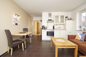 A modern kitchen with white cabinets, a stove, and a microwave. There is a dining area with a table and chairs, and a living area with a sofa and a coffee table. - Chic apartment with terrace, free swimming pool & sauna - Haus De niege Wech apartment 01 (Mönchgut)