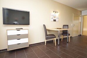 A hotel room with a flat-screen TV, a wooden cabinet, a table with two chairs, and a wall-mounted light fixture.