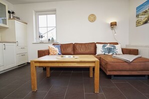 A modern living room with a brown leather sofa, a wooden coffee table, a wall clock, and a window with a view.