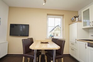 A room with a dining table, chairs, a TV, a window, and kitchen cabinets.