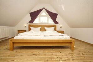 Une chambre avec un lit en bois, du linge de lit blanc, une fenêtre surmontée d’un auvent triangulaire et un radiateur.
