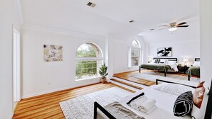 Une chambre spacieuse avec un grand lit, un plus petit lit et un ventilateur de plafond. Elle comporte deux fenêtres et offre une vue sur la verdure.