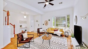 Un salon spacieux avec un grand ventilateur de plafond, un escalier, un canapé jaune, une table basse et un téléviseur.