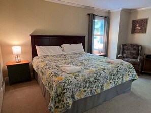 A neatly made bed with a floral bedspread, a nightstand with a lamp, a chair, and a window with blinds.