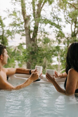 Two people in a hot tub holding white cups, surrounded by trees.