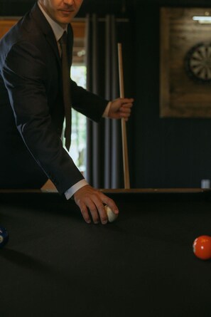 Eine Person im Anzug spielt Billard, ein Dartboard und ein Spieltisch sind zu sehen.