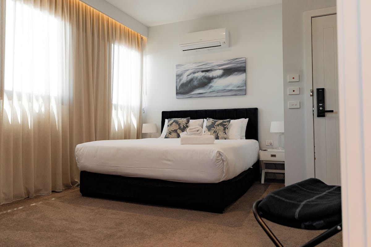 A modern hotel room with a large bed, a bedside table, a chair, and a wall-mounted air conditioner.