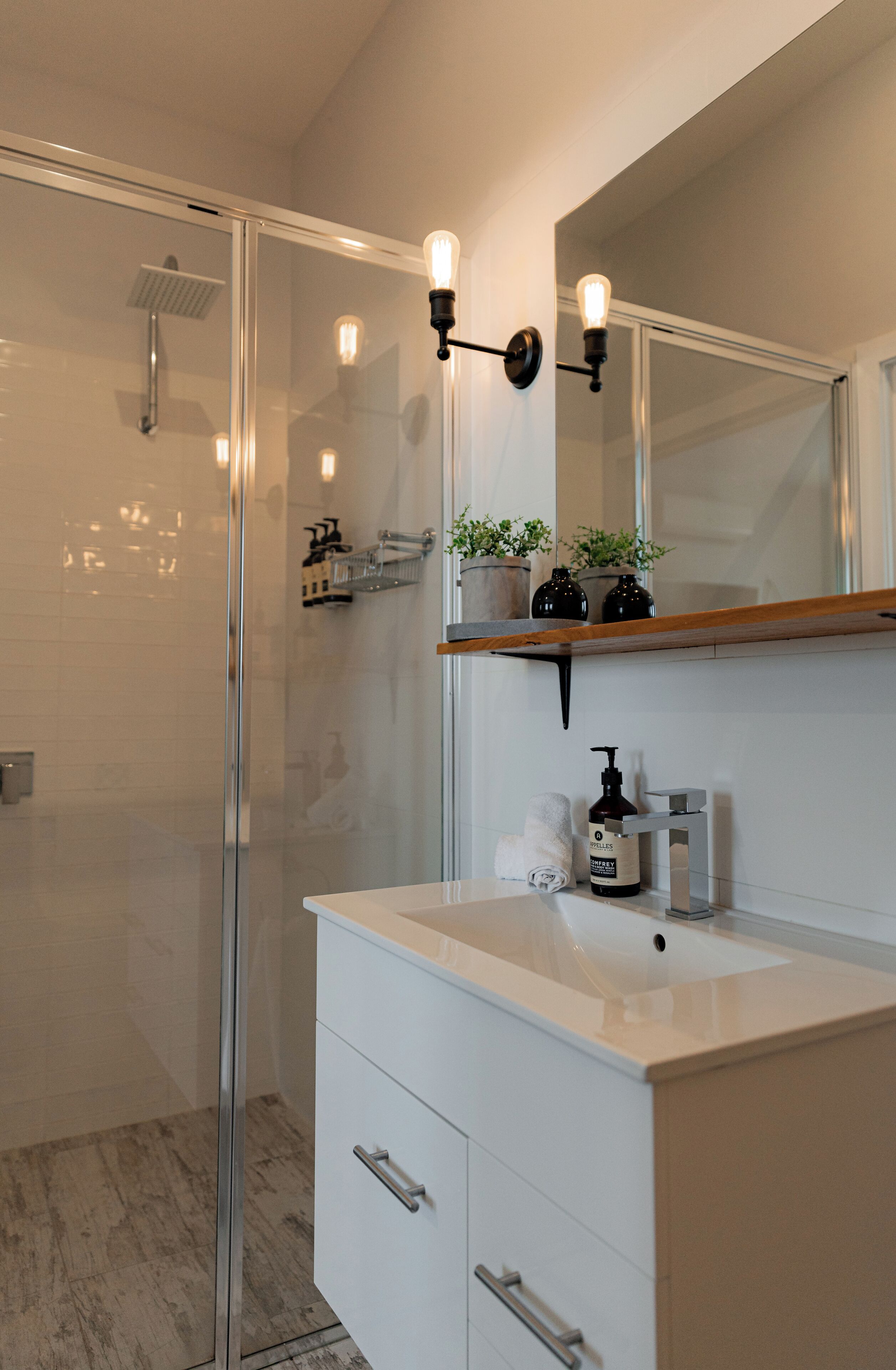 A modern bathroom with a glass shower enclosure, a white vanity with a mirror, and a shelf with toiletries.