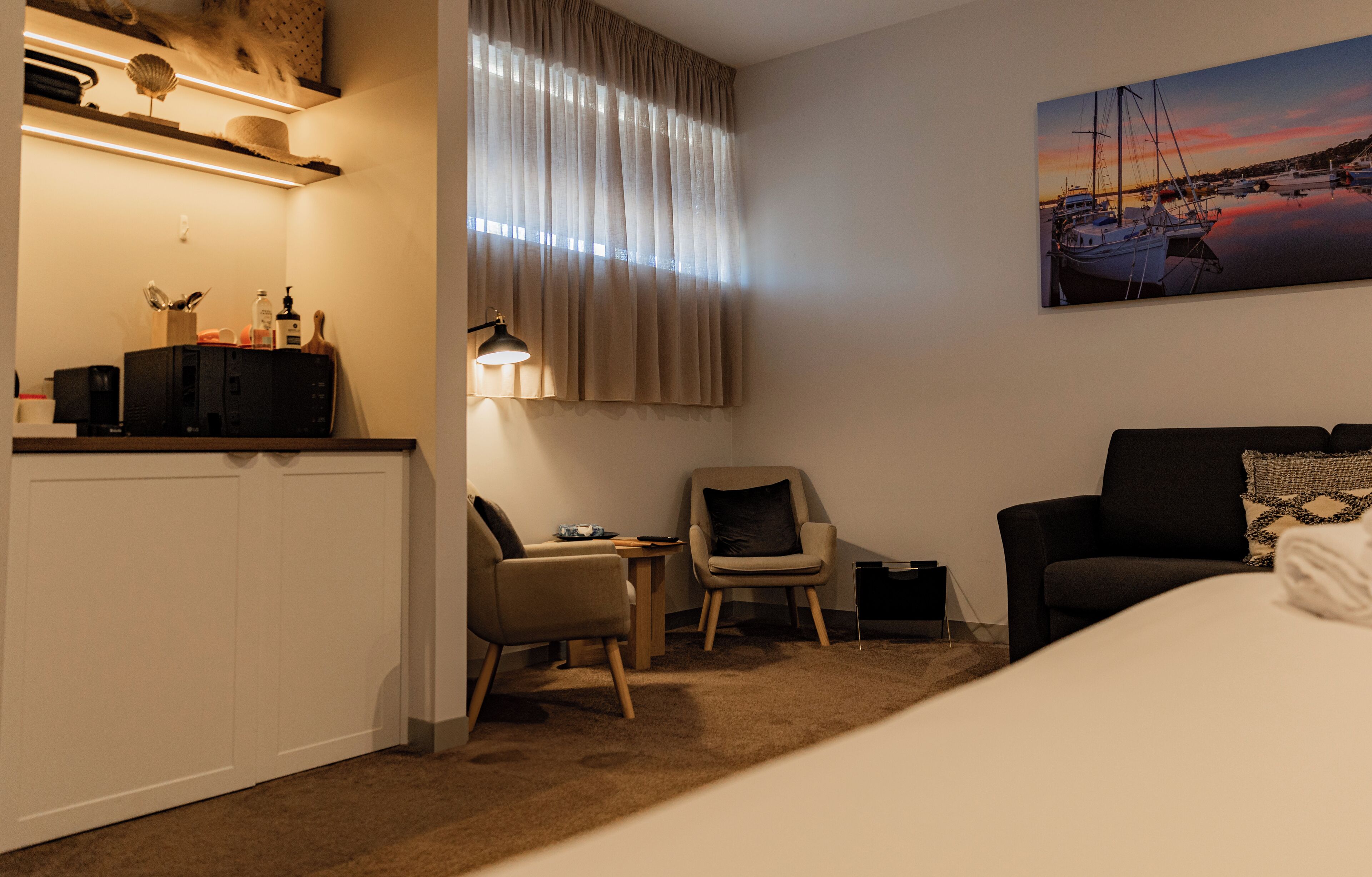 Et hotelværelse med en seng, to stole, et skrivebord, en lampe og et billede af en havn.