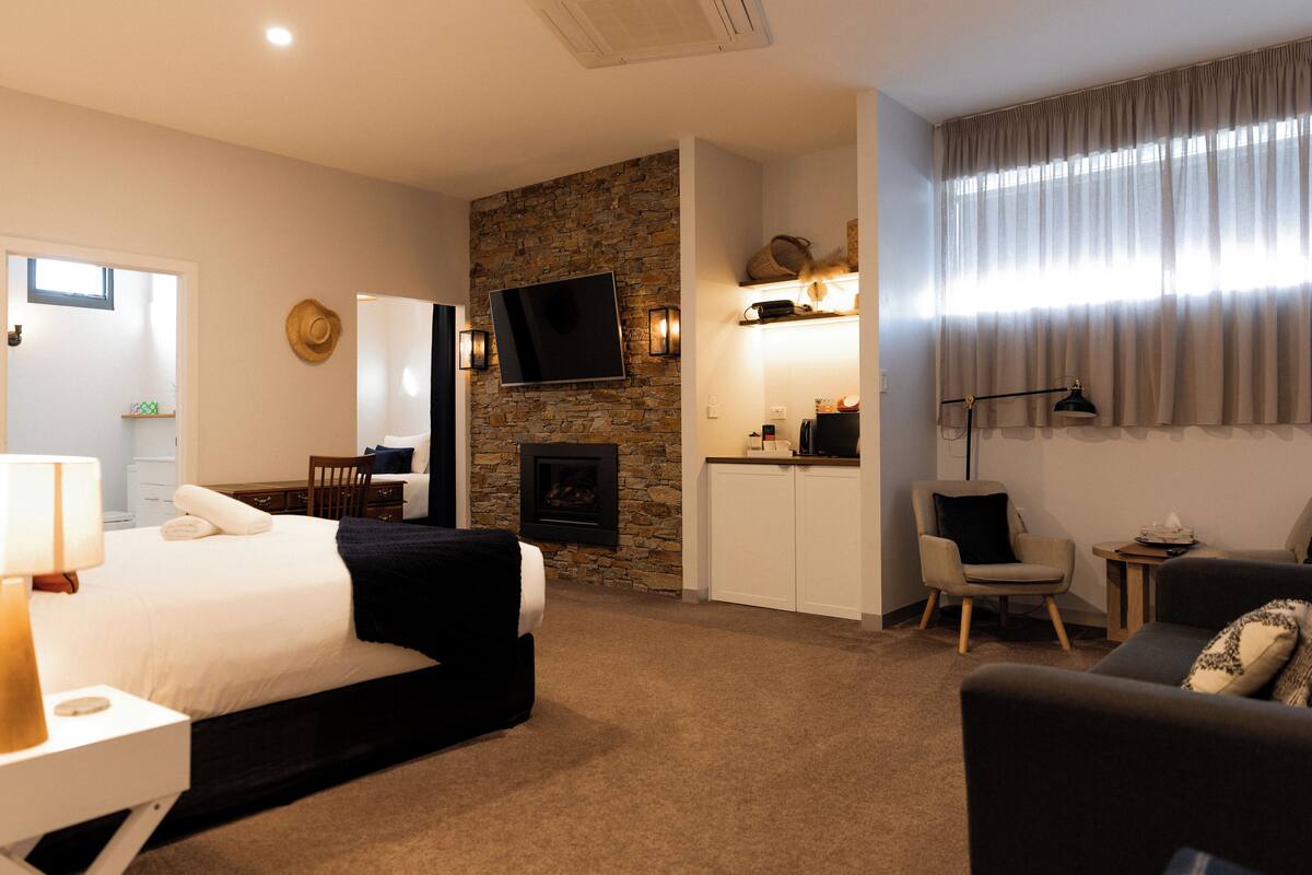 A hotel room with a bed, a fireplace, a TV, a desk with a lamp, a chair, and a sofa.