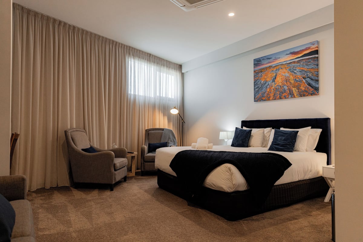 A hotel room with a large bed, two armchairs, a desk, and a painting on the wall.
