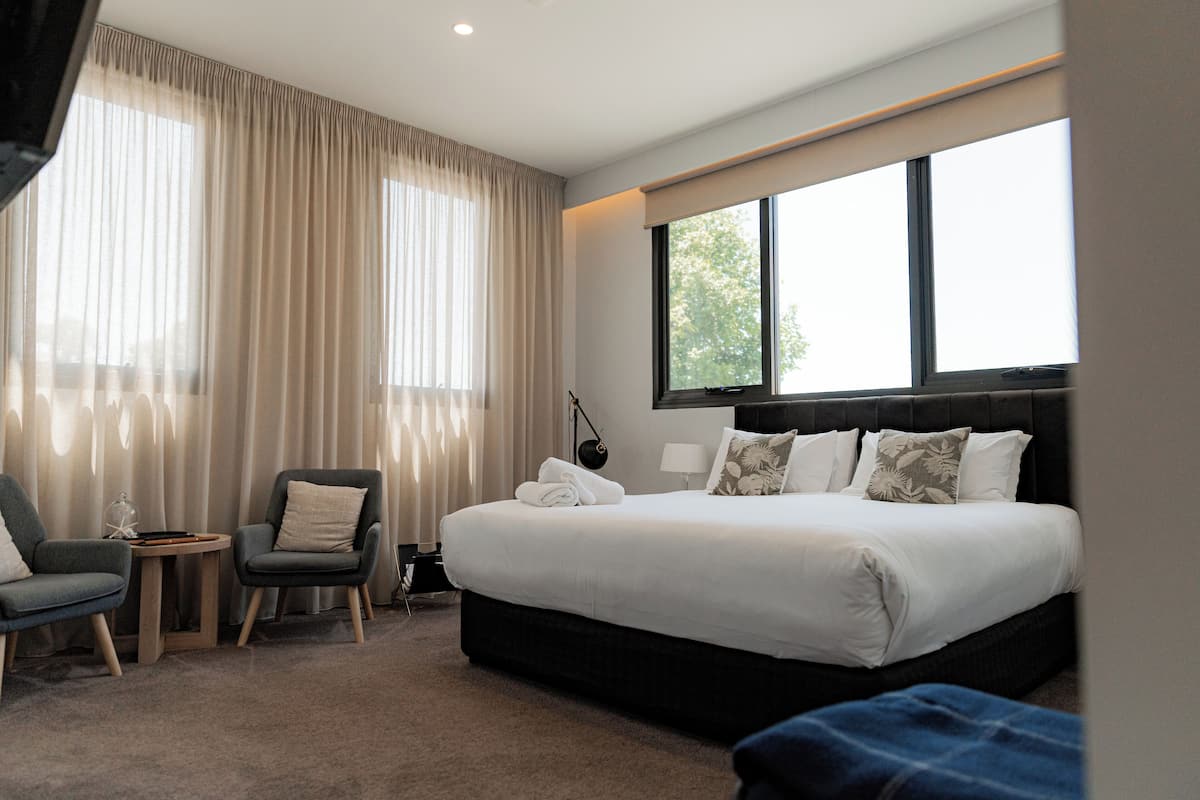 A hotel room with a large bed, two armchairs, a small side table, and a window with curtains.