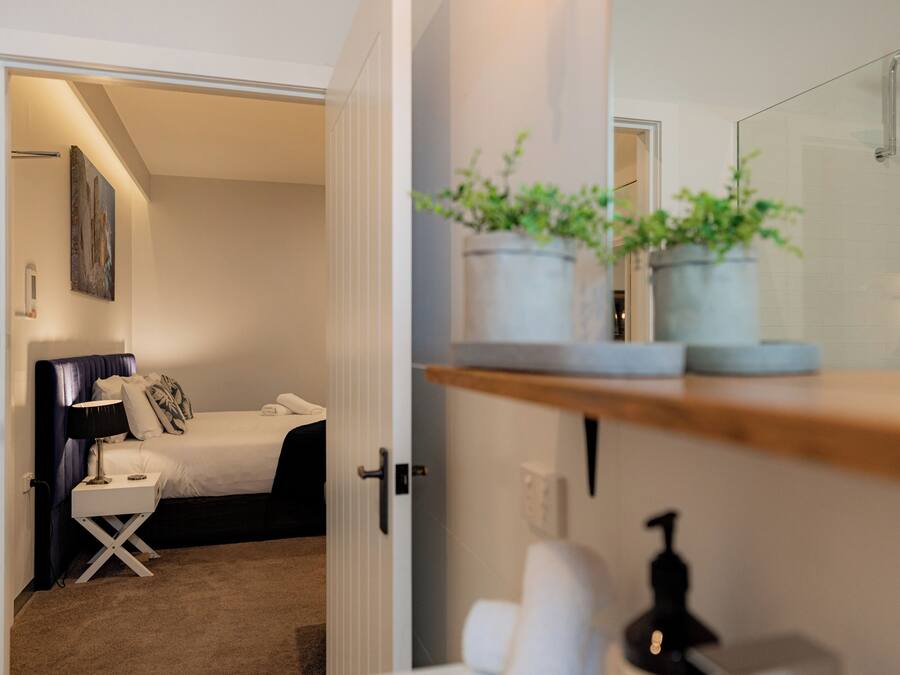 Una habitación de hotel con cama, mesita de noche, lámpara y una planta en un estante.