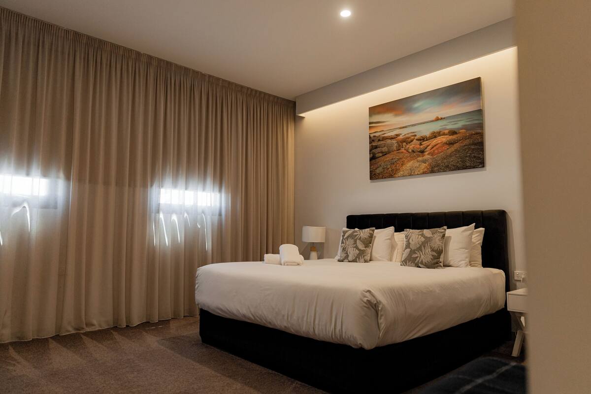 A hotel room with a large bed, a painting on the wall, and a window with curtains.