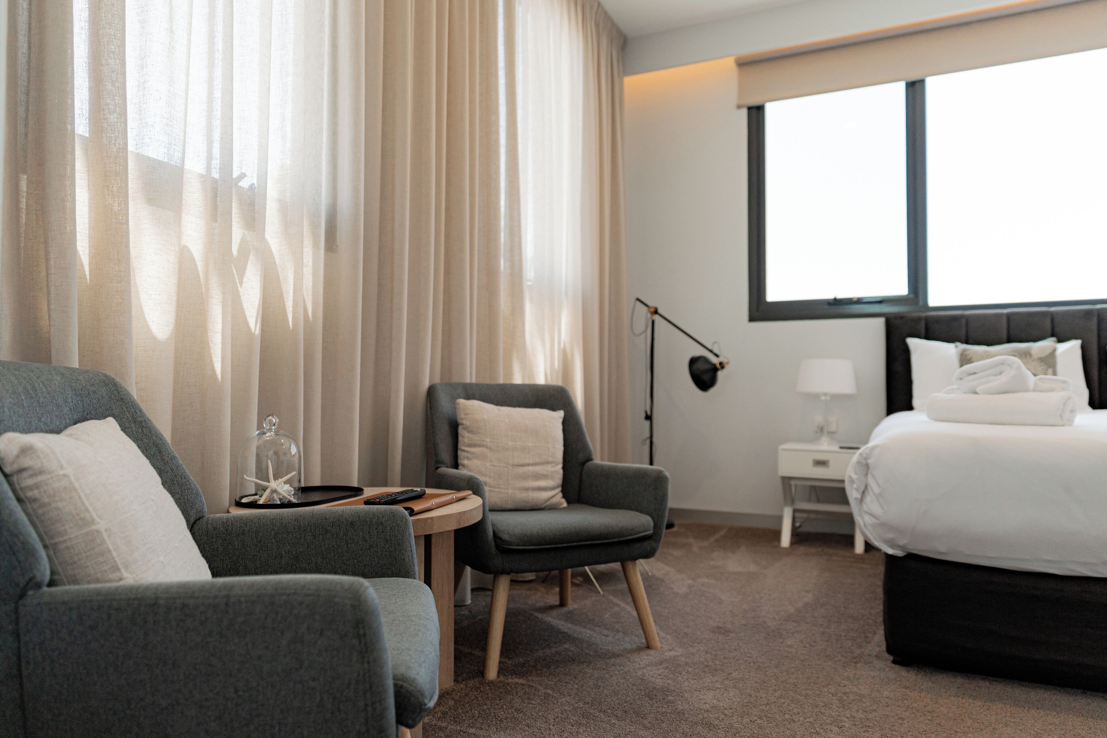 A hotel room with a bed, two armchairs, a small table, a lamp, and a large window.