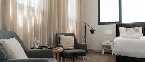A hotel room with a bed, two armchairs, a small table, a lamp, and a large window.