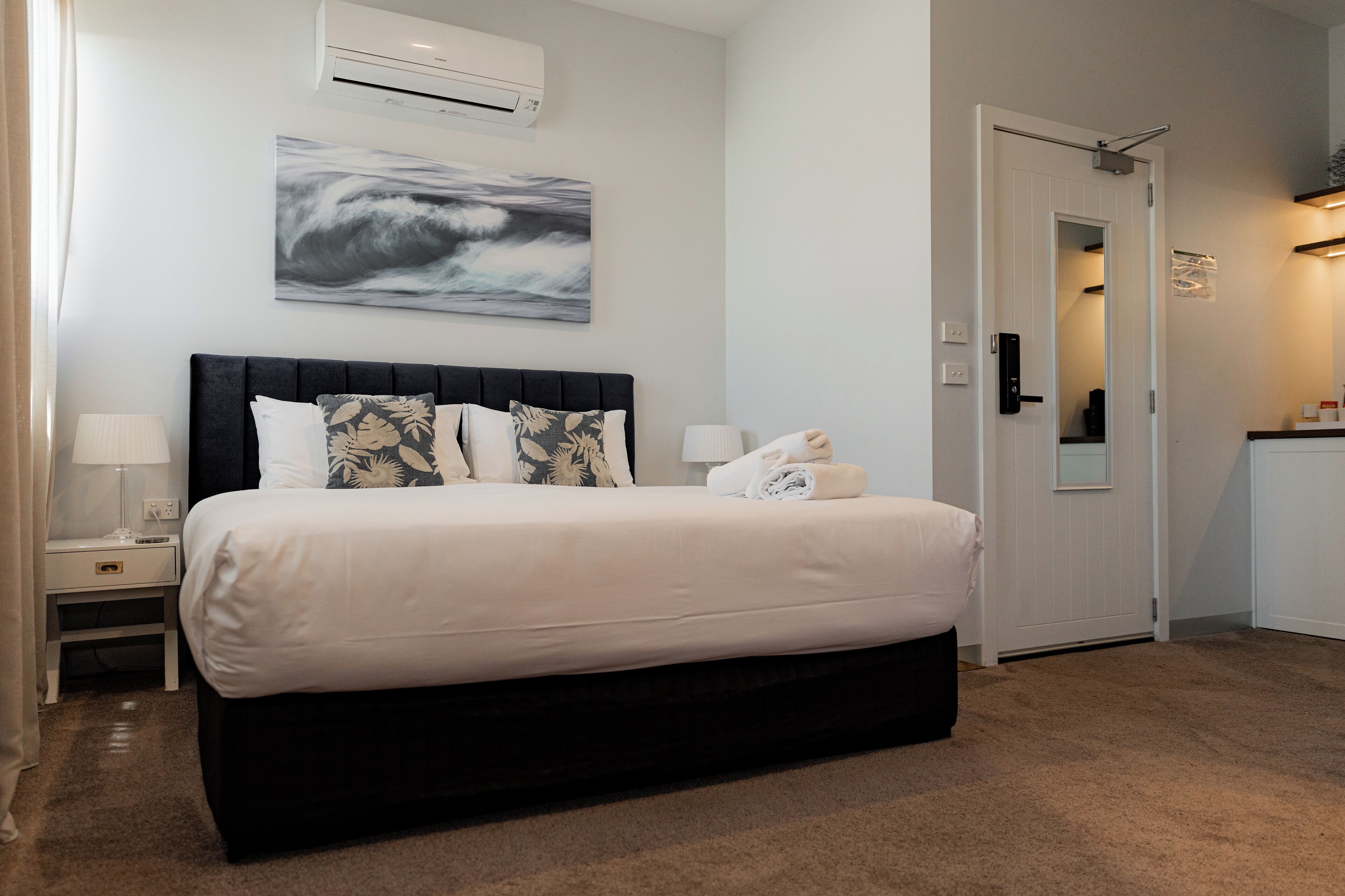 A neatly made bed with a dark headboard, a wall-mounted air conditioner, and a framed abstract painting above it.