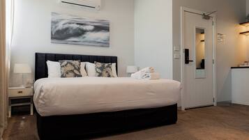 A neatly made bed with a dark headboard, a wall-mounted air conditioner, and a framed abstract painting above it.