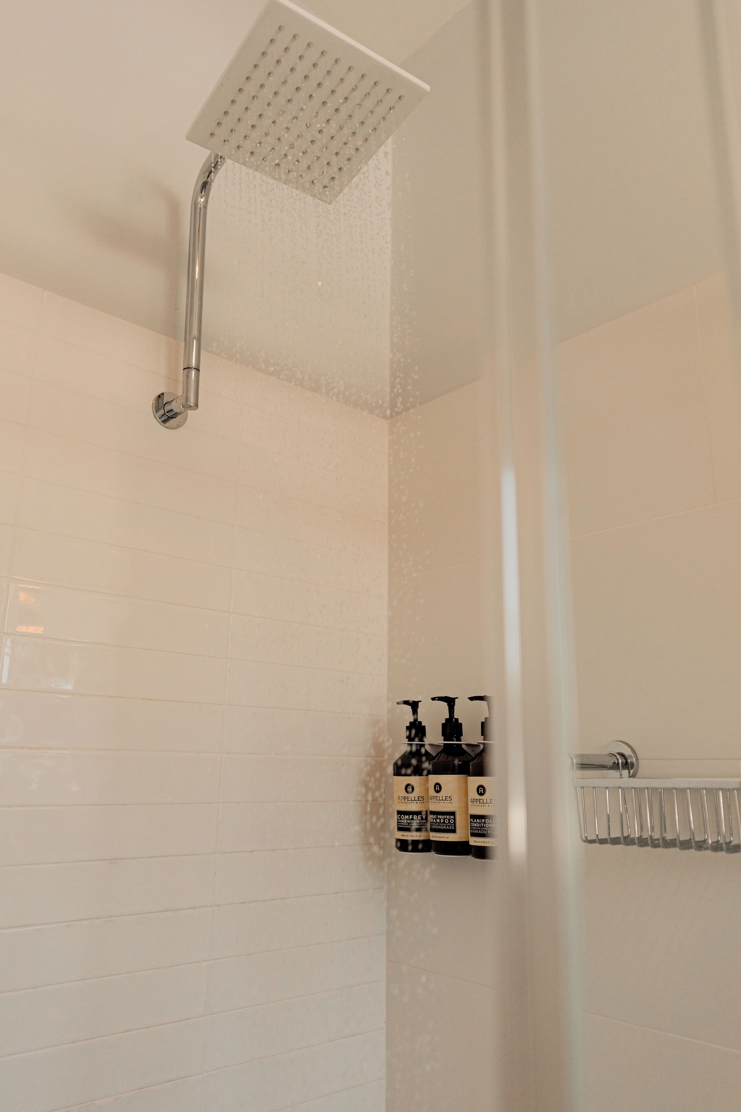 A shower with a square head, a handheld showerhead, and a wall-mounted control panel with bottles of toiletries.
