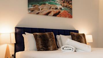 A hotel room with a bed, two bedside lamps, a wall-mounted air conditioner, and a painting of a coastal landscape.