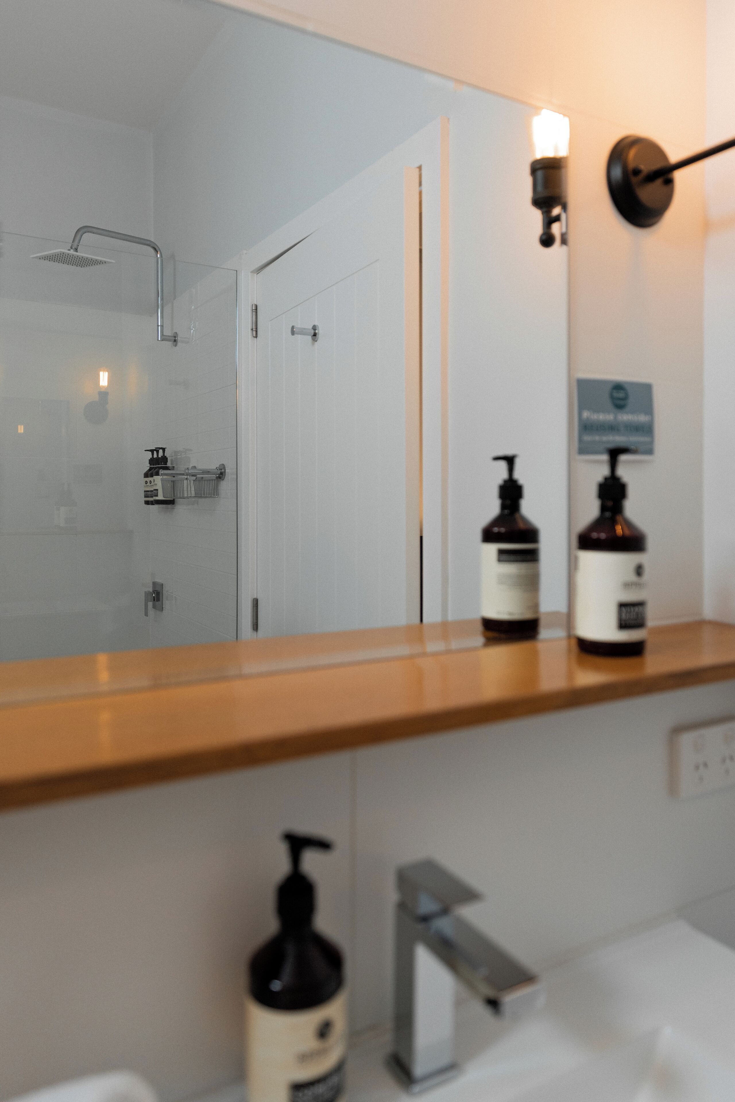 Een badkamer met een houten wastafelmeubel, een grote spiegel, een douche en twee flessen lotion op het aanrecht.
