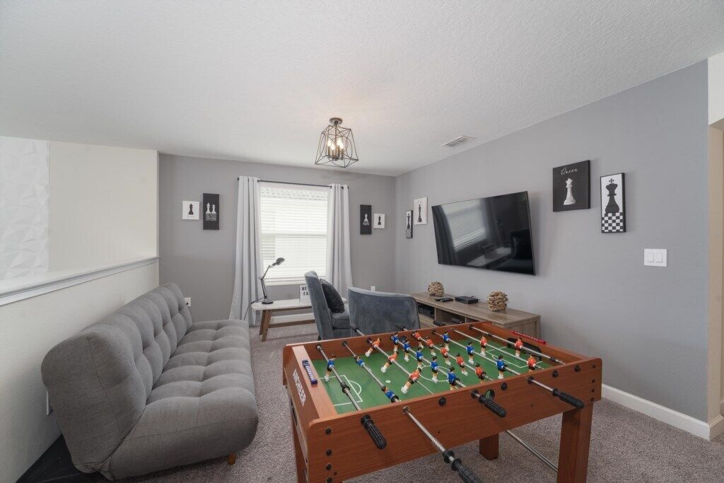 A room with a foosball table, a grey sofa, a television, and framed pictures on the wall.