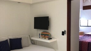 Una habitación de hotel con una cama, un televisor montado en la pared, un estante pequeño con una botella y un control remoto, y una puerta que da a otra habitación.