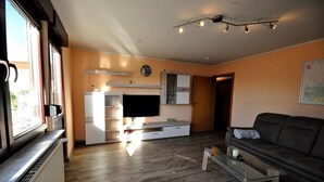 Een moderne woonkamer met een bank, salontafel en televisie. Er is een keukenhoek met kasten en een raam.