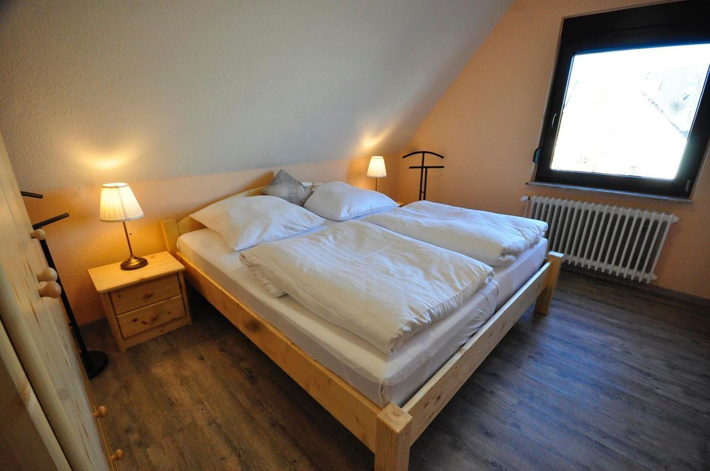 Un dormitorio con una cama de madera, dos mesitas de noche con lámparas, un radiador y una ventana con vistas.