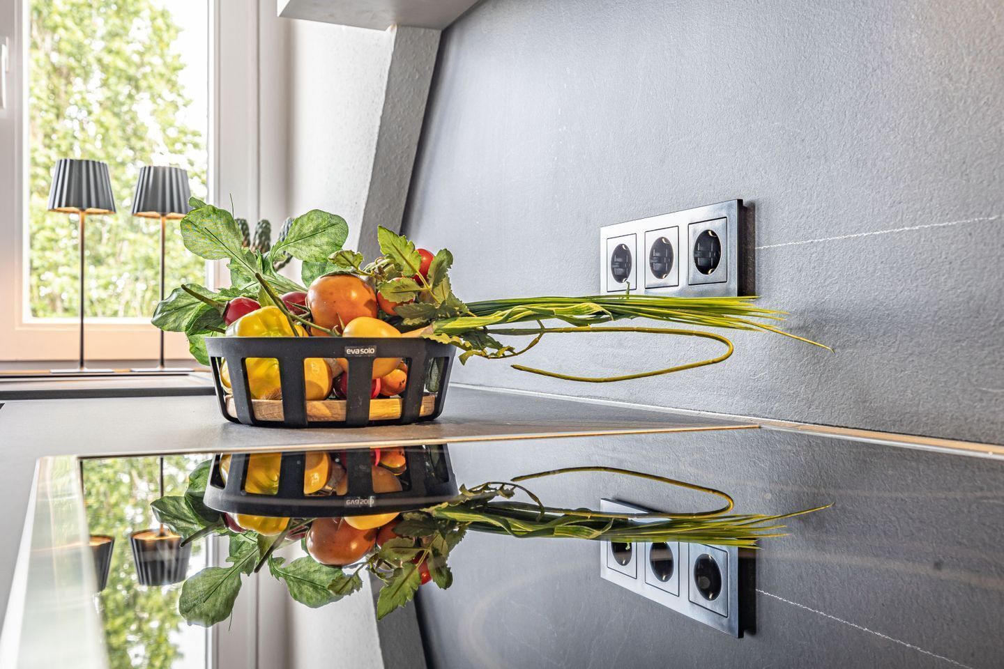 Een keuken met een mand met fruit in het zwart op het aanrecht, een raam met uitzicht op bomen, en een wandcontactdoos.