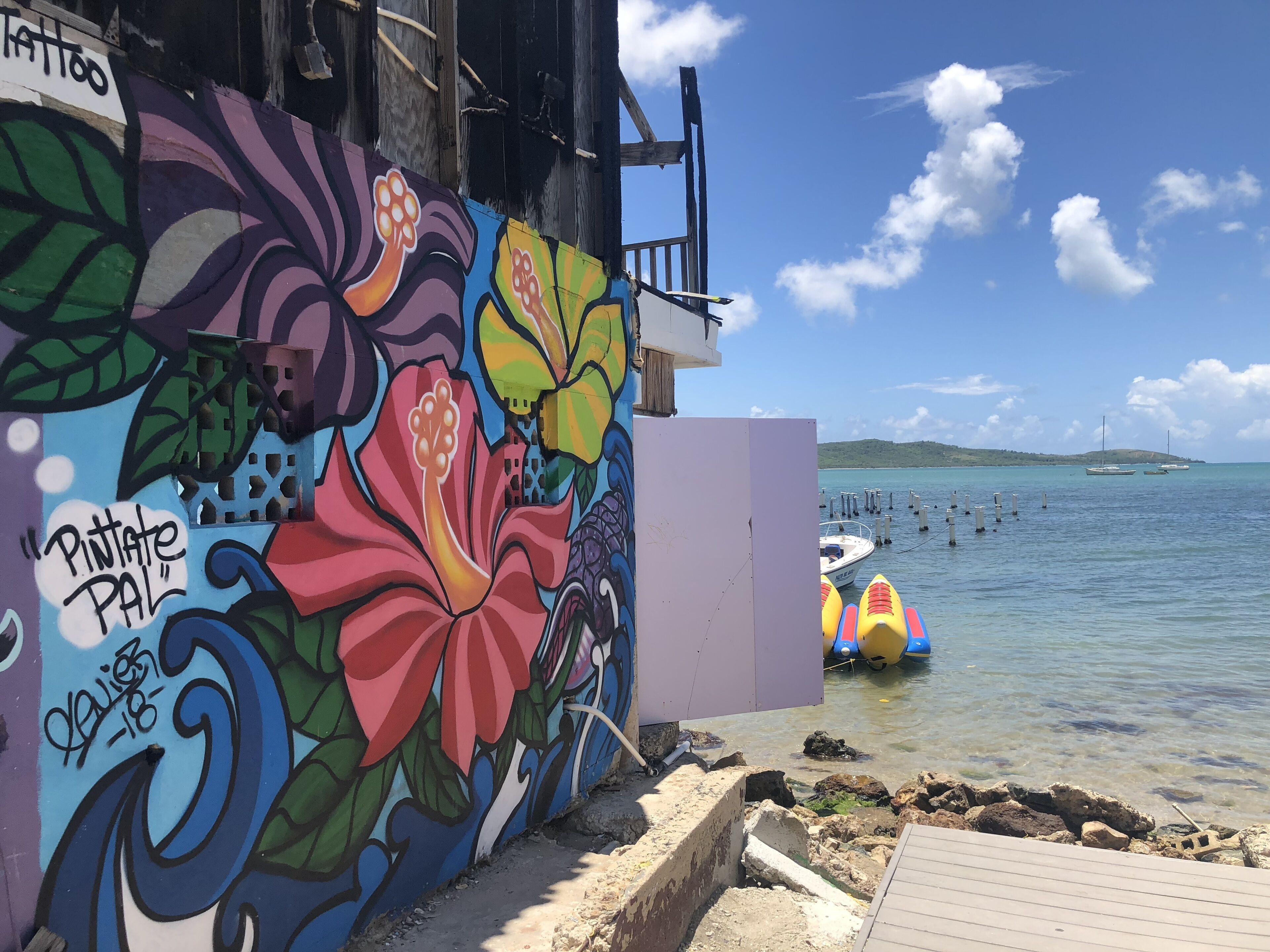 A colorful mural with floral designs and graffiti, a wooden dock, and a calm sea with boats.