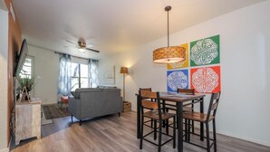 A modern living room with a grey sofa, wooden dining table with four chairs, a ceiling fan, and a wall with colorful geometric art. - Condo in the heart of the Catalina Foothills (Tucson)