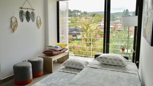 Ein Schlafzimmer mit großem Fenster, einem Bett mit gemusterten Kissen, einer Bank mit Kissen und an der Wand befestigten Traumfängern.